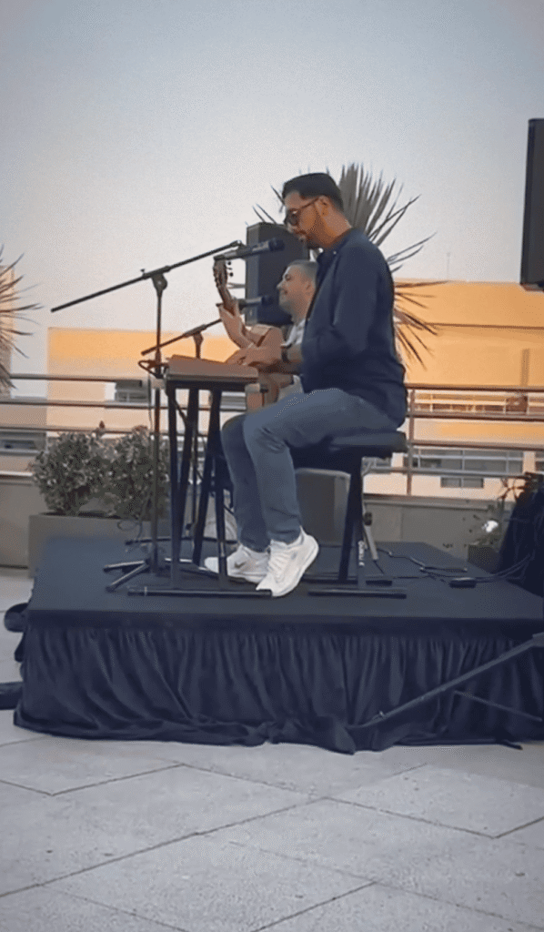 Dois músicos num pequeno palco ao ar livre, um sentado a cantar ao microfone enquanto toca um cajón e outro ao fundo com uma guitarra, com colunas de som e plantas atrás; ambiente de festa, ideal para um trio de pop espanhol e rumba para festas maiores em Barcelona.
