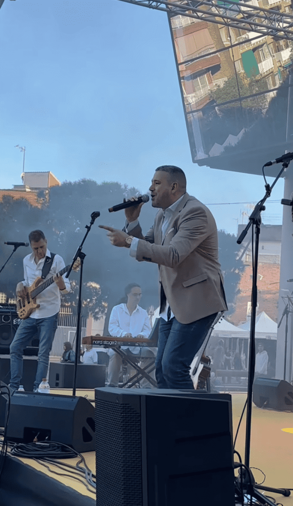 Num palco ao ar livre, um cantor de blazer bege canta ao microfone e aponta para o público, enquanto um baixista à esquerda e um teclista ao fundo acompanham a música; ao fundo vê se um ecrã grande com prédios refletidos, sugerindo um evento de rua, ideal para quem procura um trio de pop espanhol e rumba para festas maiores em Barcelona.