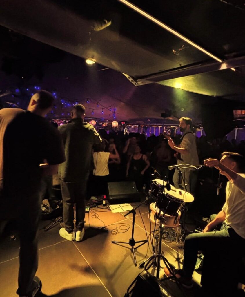 Palco pequeno num espaço interior com luz baixa e cordão de luzes azuis ao fundo, com músicos a tocar para um público muito próximo; vê se um baterista sentado à direita junto a um kit de bateria e um guitarrista de pé, enquanto a plateia dança e levanta os braços, num ambiente de festa que combina rumba fusão e flamenquito para um aniversário de verão em Barcelona.