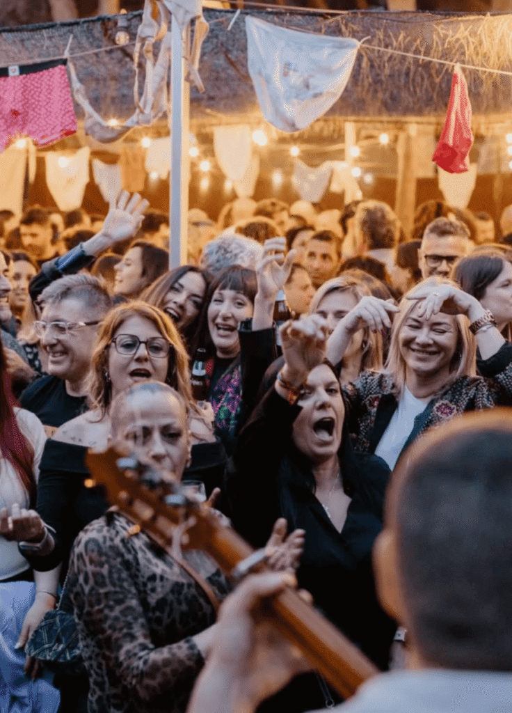 Multidão a dançar e a cantar num evento ao ar livre ao entardecer, com luzes suspensas e roupa pendurada por cima; em primeiro plano vê se a guitarra desfocada de um músico, enquanto o público sorri e levanta as mãos, num ambiente festivo em Barcelona, ideal para um trio de pop espanhol e rumba para festas maiores.