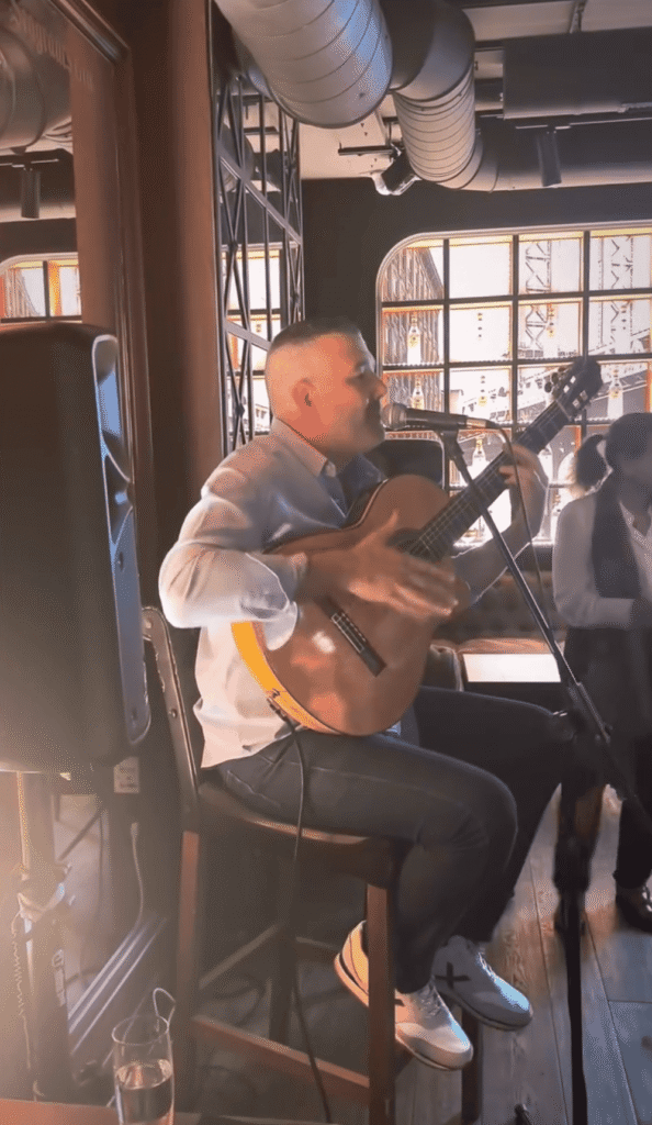 Ein Musiker sitzt auf einem Barhocker in einer Innenlocation und spielt eine klassische Gitarre, während er in ein Mikrofon singt. Neben ihm stehen Lautsprecher und Kabel, im Hintergrund sieht man große Fenster und sichtbare Lüftungsrohre an der Decke. Die Szene passt zu einem Trio aus spanischem Pop und spanischer Rumba für Festtage in Barcelona, geeignet für Hochzeiten und private Feiern.