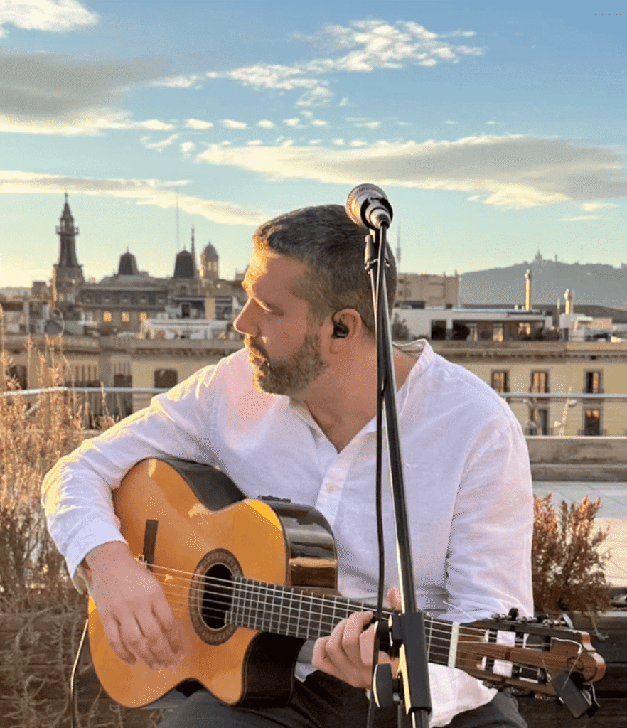 Ein Musiker in einem weißen Hemd sitzt auf einer Dachterrasse und hält eine klassische Gitarre in Spielposition. Vor ihm steht ein Mikrofonständer, im Ohr trägt er einen In Ear Monitor. Im Hintergrund sieht man die Dächer und Türme von Barcelona bei warmem Abendlicht, passend als Bild für ein Trío de pop español y rumba für Fiestas Mayores in Barcelona.