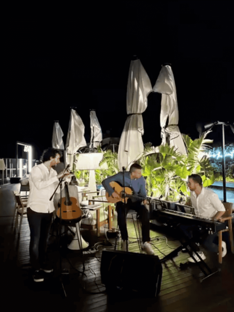 Nachts auf einer Terrassenlocation spielen drei Musiker spanische Popmusik und spanische Rumba: links steht ein Sänger am Mikrofon, in der Mitte sitzt ein Gitarrist auf einem Hocker, rechts spielt ein Pianist am Keyboard. Hinter ihnen sind geschlossene Sonnenschirme, Pflanzen und eine beleuchtete Pool oder Gartenanlage zu sehen. Die Szene passt zu einem Trio für private Feiern und Stadtfeste in Barcelona.