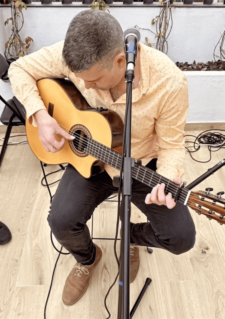 Ein Gitarrist sitzt auf einem Stuhl und spielt eine spanische Akustikgitarre, während ein Mikrofonständer direkt vor ihm steht. Im Hintergrund sieht man eine helle Wand mit Pflanzen und auf dem Boden liegen Kabel, was auf einen kleinen Aufbau für eine Feier hindeutet. Passend für ein Trio aus spanischem Pop und spanischer Rumba für Stadtteilfeste in Barcelona.