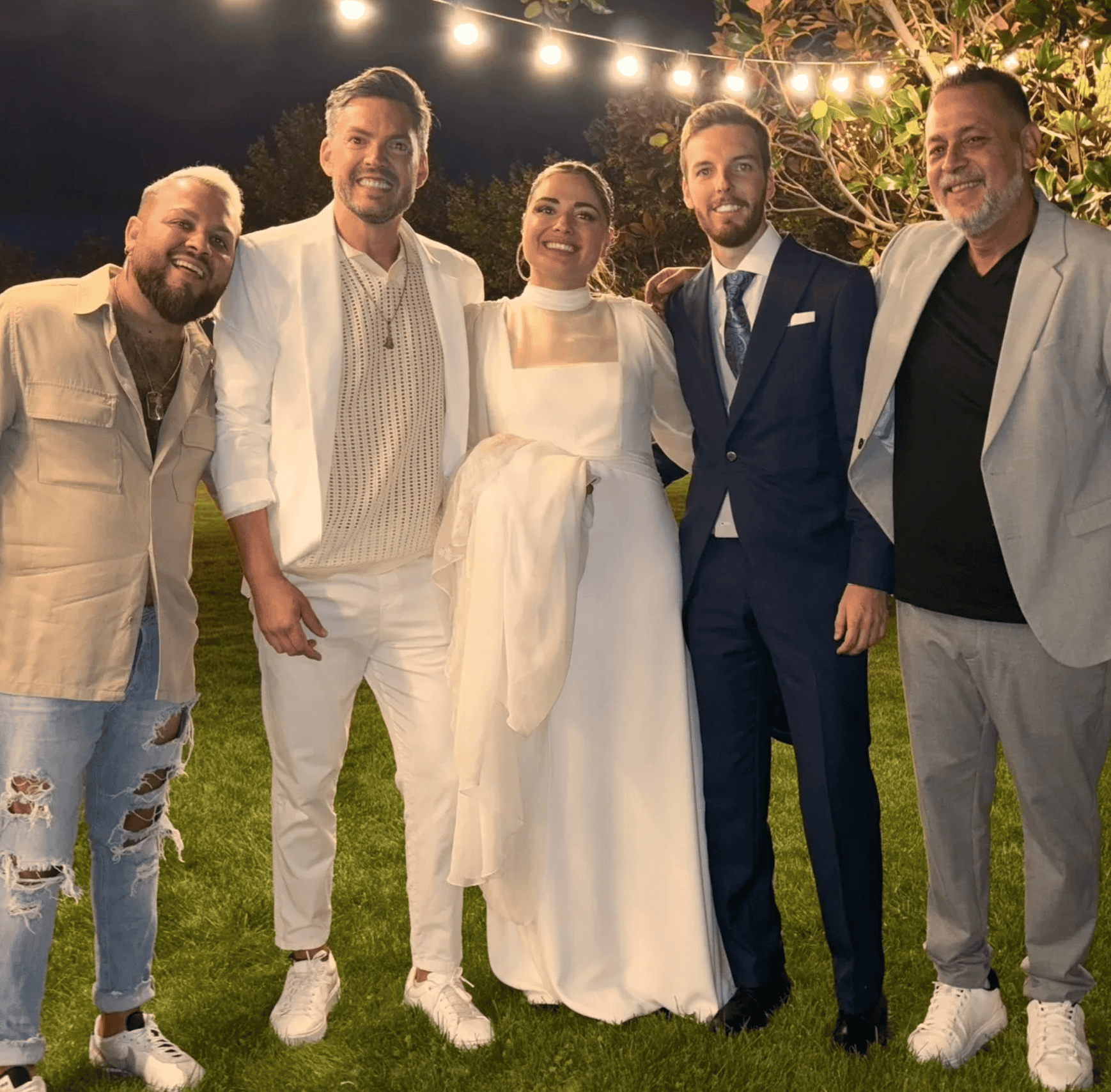 夜の屋外の芝生で、電球のガーランドの下に並んで立つ五人の集合写真。中央に白いドレスの新婦、その右に紺のスーツの新郎。両側にカジュアルな服装の男性が寄り添い、笑顔でカメラを見ている。マドリードで結婚式のパーティー向けにスペインの祝祭音楽を届ける二人組の融合系グループの雰囲気を伝えるカット。