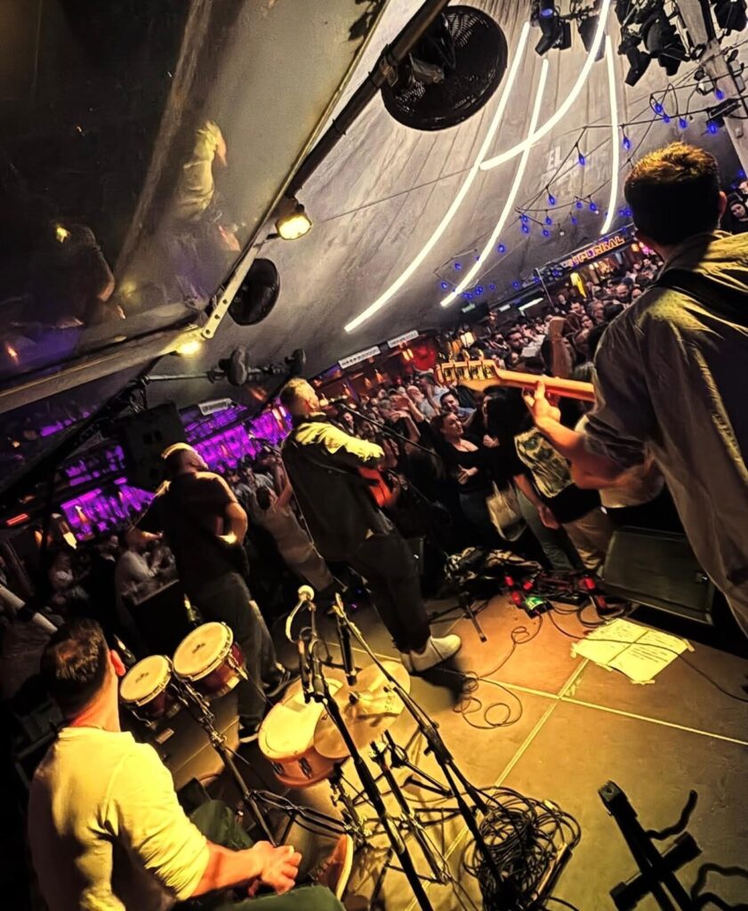 Foto tirada do palco num espaço interior cheio, com uma banda a tocar para uma multidão. À direita vê se um músico de costas com guitarra elétrica e, ao centro, outro com violão junto ao microfone. À esquerda aparecem congas e cabos no chão do palco. No teto há arcos de luz e pequenas lâmpadas azuis, criando ambiente de festa, ideal para rumba fusão e flamenquito num aniversário de verão em Barcelona.