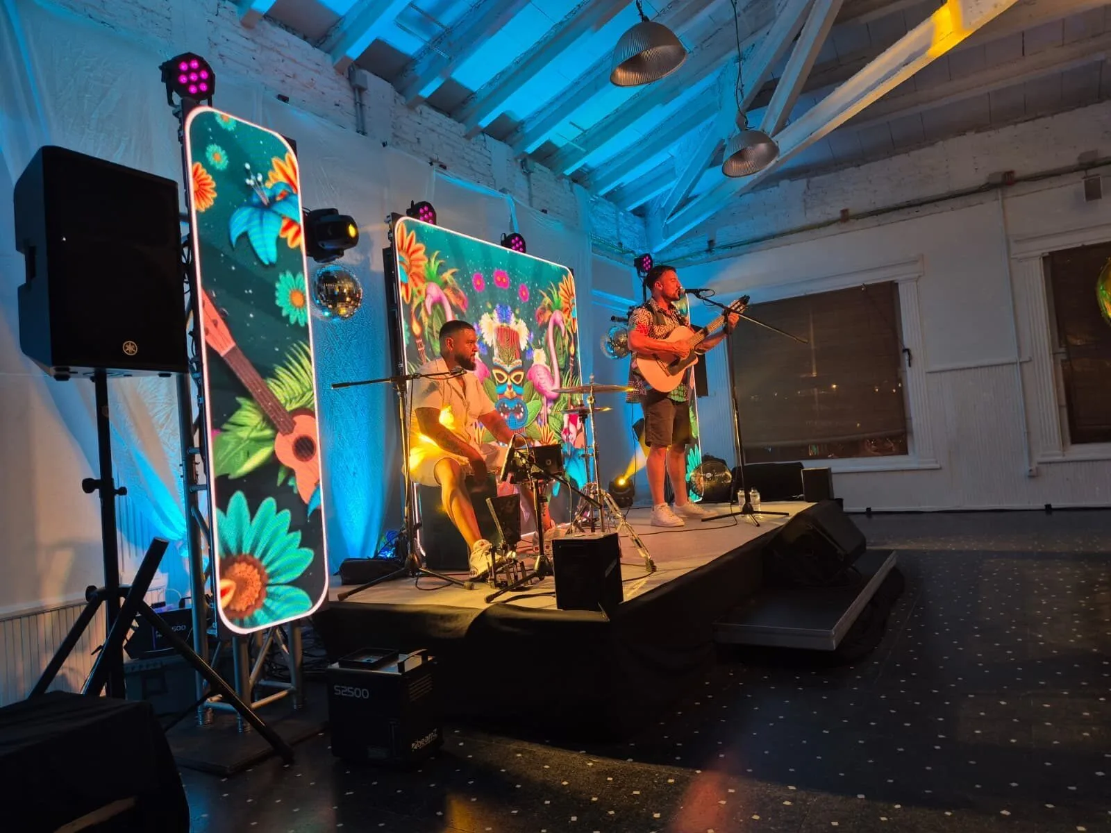 Dos músicos sobre un escenario en un salón interior: a la derecha, un cantante toca una guitarra y canta en un micrófono; a la izquierda, un percusionista sentado toca un cajón. Detrás hay paneles luminosos con ilustraciones tropicales y luces de escenario, creando un ambiente de rumba fusión en Barcelona para bodas con ganas de fiesta.