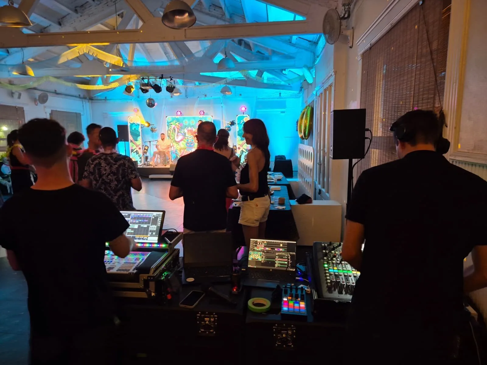 Vista desde la mesa de control de sonido en una sala de eventos: dos técnicos manejan una mesa de mezclas y un portátil mientras, al fondo, un pequeño escenario iluminado en azul y rosa recibe a los músicos y varias personas conversan y se mueven cerca de la pista. Ambiente de rumba fusión en Barcelona para bodas con ganas de fiesta.