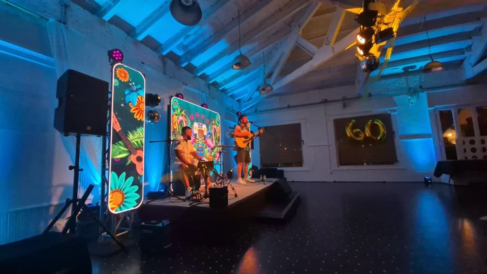 Dos músicos actúan sobre un pequeño escenario en una sala interior: uno canta y toca la guitarra con un micrófono, y el otro está sentado con un instrumento de percusión. Detrás hay paneles decorativos con dibujos tropicales iluminados con luces de colores y, a la derecha, se ven globos dorados con el número 60. La escena transmite rumba fusión en Barcelona para bodas y celebraciones con ganas de fiesta.