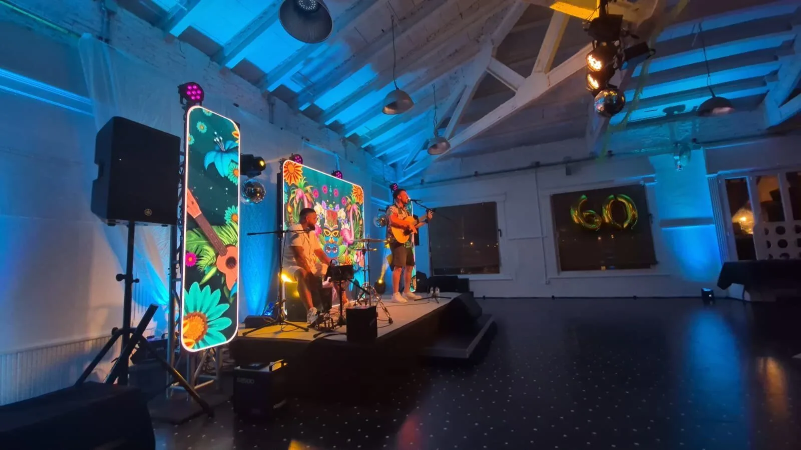 Escenario interior con dos músicos actuando: uno canta y toca la guitarra de pie ante un micrófono y otro está sentado tocando un cajón y percusión. La sala está iluminada en azul y en el fondo se ven dos paneles decorativos con flores y calaveras de colores, además de globos dorados con el número 60, en un ambiente de rumba fusión en Barcelona para bodas con ganas de fiesta.