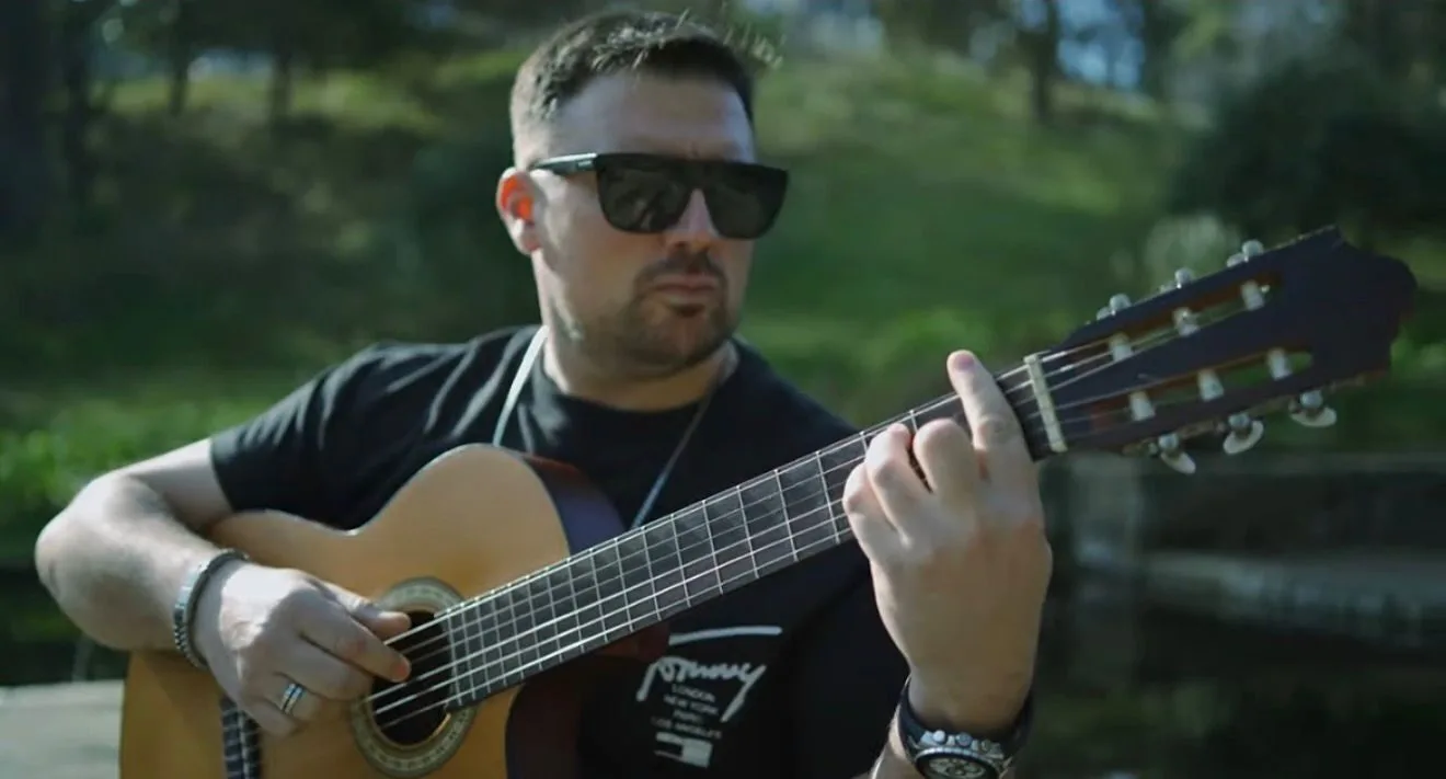 Primer plano de un guitarrista con gafas de sol tocando una guitarra española al aire libre, con un fondo verde desenfocado; imagen pensada para promocionar rumba fusión en Barcelona para bodas y celebraciones con ganas de fiesta.