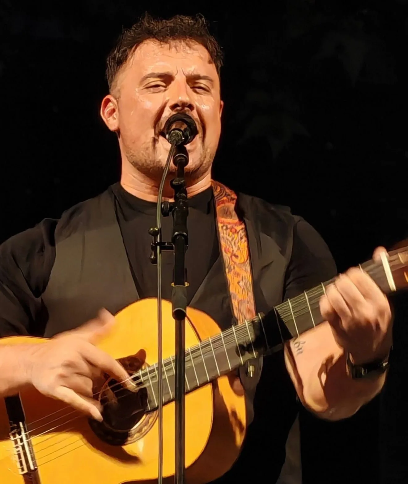 Primer plano de un cantante con camiseta negra cantando al micrófono mientras toca una guitarra española; se ve la correa estampada y el fondo oscuro de escenario, una imagen que encaja con una rumba fusión en Barcelona para bodas con ganas de fiesta.