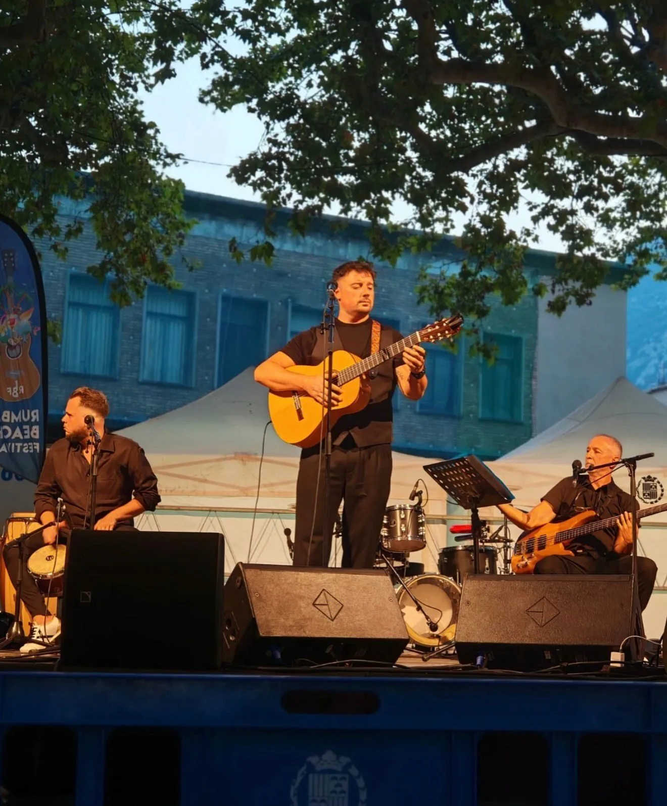 Tres músicos sobre un escenario al aire libre: en el centro, un guitarrista de pie junto a un micrófono; a la izquierda, un percusionista sentado con un cajón; a la derecha, un bajista sentado con su instrumento. Hay monitores en el suelo, una batería al fondo y ramas de árboles sobre el escenario. Imagen asociada a rumba fusión en Barcelona para bodas con ganas de fiesta.