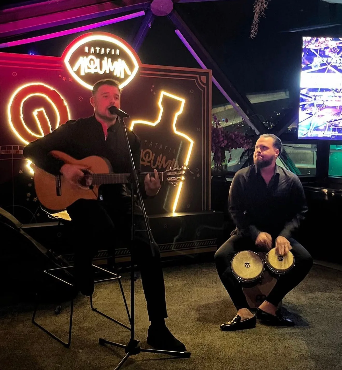 Dos músicos actuando en un interior con luces de neón: a la izquierda, un cantante toca la guitarra sentado y canta con un micrófono; a la derecha, otro músico sentado toca unos bongos. La escena transmite ambiente de rumba fusión en Barcelona para bodas con ganas de fiesta.
