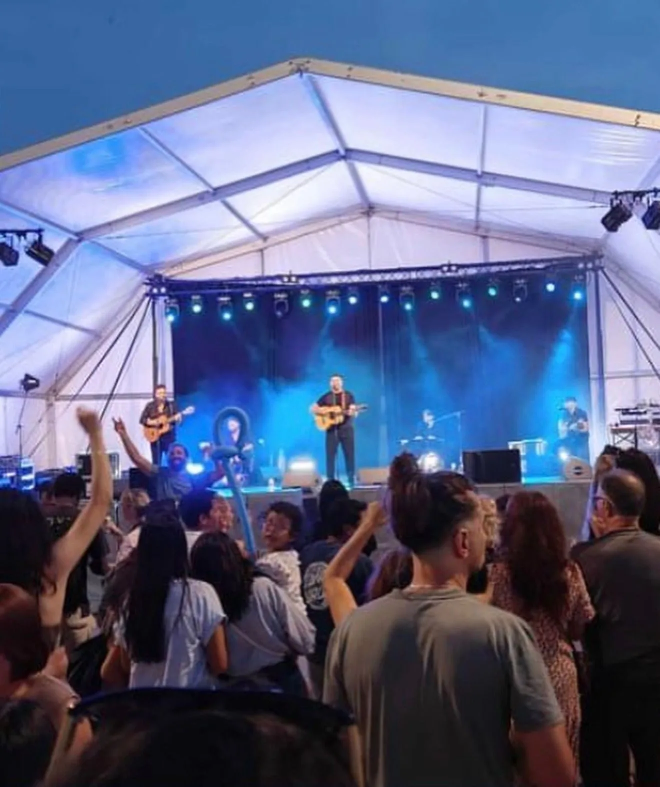 Público con los brazos en alto frente a un escenario bajo una carpa blanca; varios músicos tocan guitarras con iluminación azul, creando un ambiente de rumba fusión en Barcelona para bodas con ganas de fiesta.