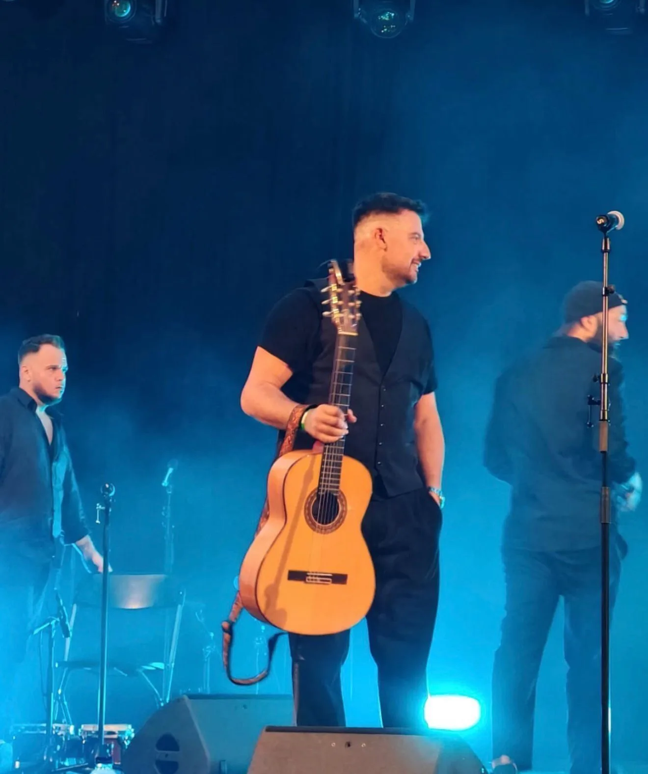 En un escenario con luz azul y algo de humo, un músico sonríe mientras sostiene una guitarra española colgada del hombro; a su lado se ve un pie de micro y, al fondo, otros integrantes preparando el momento. Imagen asociada a rumba fusión en Barcelona para bodas con ganas de fiesta.
