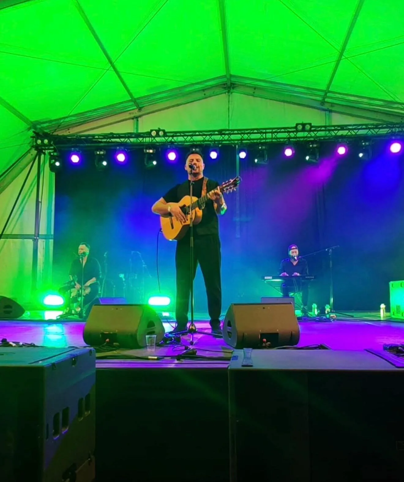 En un escenario bajo una carpa, un cantante con guitarra está en el centro frente a un micrófono, con luces verdes y moradas iluminando el fondo; a los lados se ven otros dos músicos, uno sentado y otro en un teclado. Imagen asociada a rumba fusión en Barcelona para bodas con ganas de fiesta.