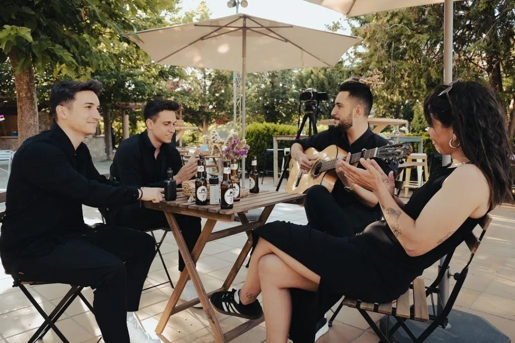 Cuatro personas sentadas alrededor de una mesa de madera en una terraza con sombrillas y árboles; un hombre toca una guitarra española mientras una mujer aplaude, en un ambiente relajado que encaja con pop español con flamenquito para bodas en Barcelona.