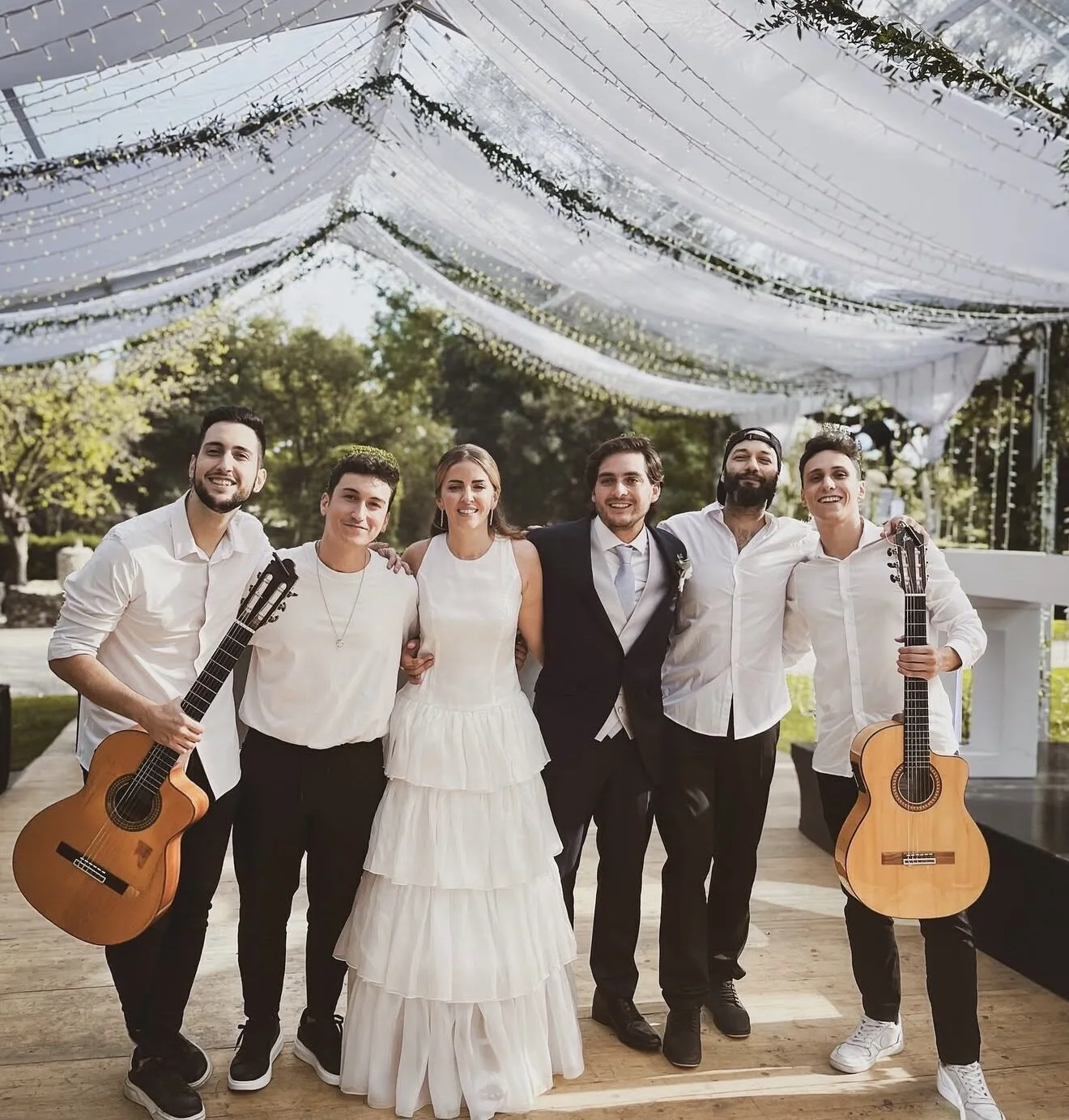 Seis personas posan juntas en una boda al aire libre: la novia con vestido blanco y el novio con traje oscuro, rodeados por cuatro músicos; dos de ellos sostienen guitarras españolas. Sobre sus cabezas hay telas blancas y guirnaldas de luces, creando un ambiente festivo, ideal para pop español con flamenquito para bodas en Barcelona.