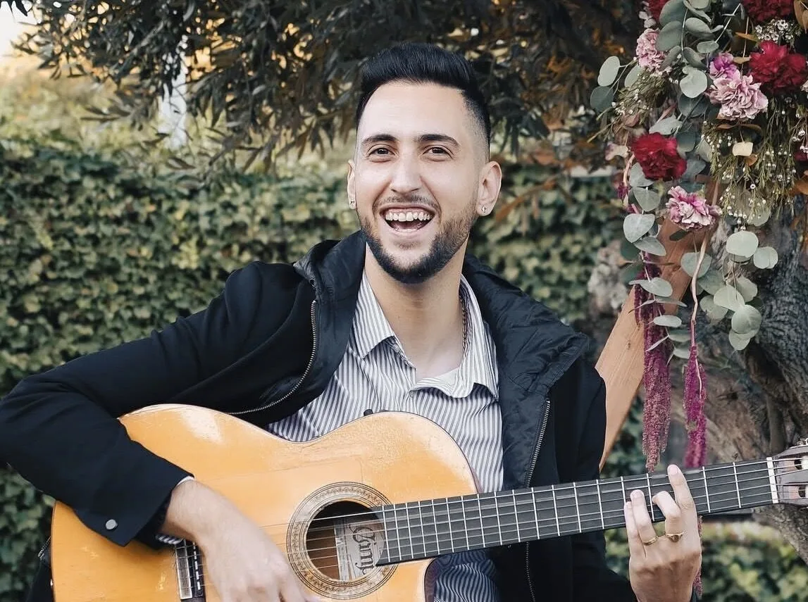Retrato de un guitarrista sonriendo mientras sostiene una guitarra española, en un entorno exterior con vegetación y una decoración floral a un lado; imagen asociada a pop español con flamenquito para bodas en Barcelona.