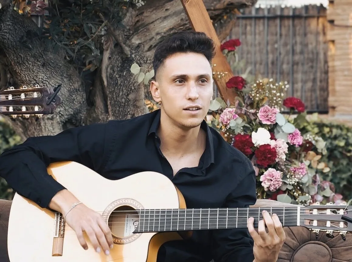 Primer plano de un guitarrista con camisa negra tocando una guitarra española, sentado al aire libre junto a un árbol y una decoración de flores de colores; imagen adecuada para pop español con flamenquito para bodas en Barcelona.