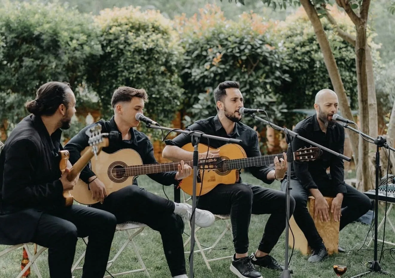 Cuatro músicos vestidos de negro sentados en un jardín, tocando guitarras españolas y cantando con micrófonos; uno acompaña con un cajón. La escena muestra un formato acústico de pop español con flamenquito, ideal para bodas en Barcelona.