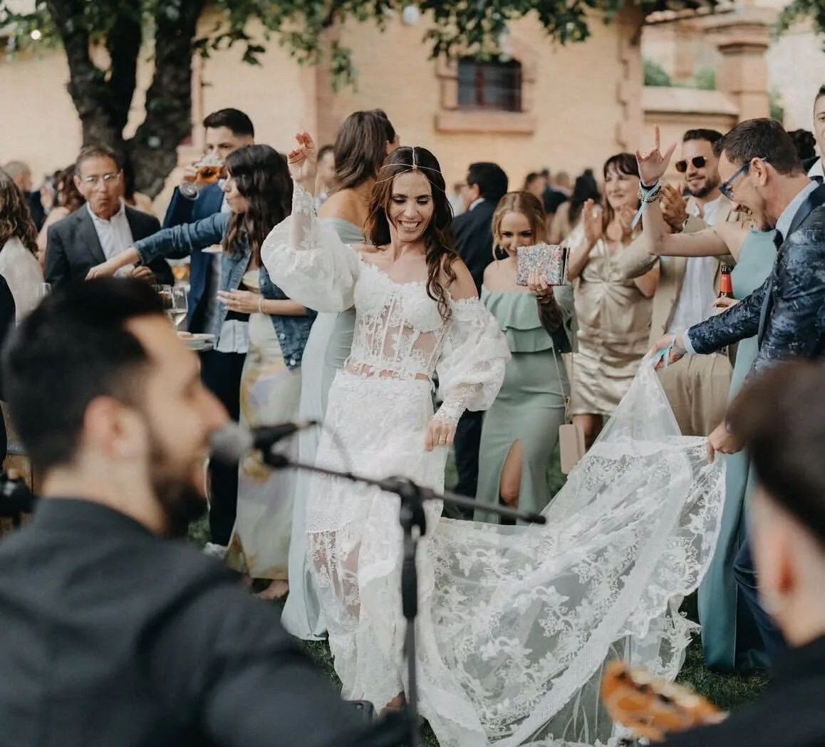 Novia con vestido blanco de encaje bailando sonriente en un jardín durante una boda, rodeada de invitados que aplauden; en primer plano se ven desenfocados un micrófono y música, ambiente ideal para pop español con flamenquito para bodas en Barcelona.