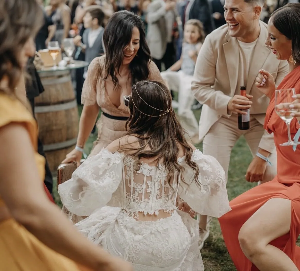 Invitados bailando y riendo en una boda al aire libre; una mujer con vestido blanco de encaje se mueve en el centro mientras a su alrededor varias personas brindan con copas y una botella, creando un ambiente festivo ideal para pop español con flamenquito para bodas en Barcelona.