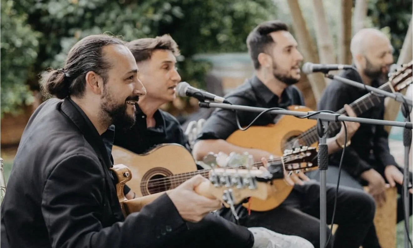 Cuatro músicos sentados en fila, vestidos de negro, cantando a micrófonos y tocando guitarras españolas en un espacio exterior con árboles al fondo; imagen típica de pop español con flamenquito para bodas en Barcelona.