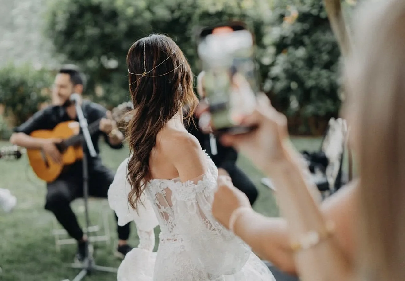De espaldas, una novia con vestido blanco de encaje y el pelo suelto mira hacia un músico con guitarra y un micrófono en un jardín; en primer plano, una invitada levanta una copa. Imagen de pop español con flamenquito para bodas en Barcelona.