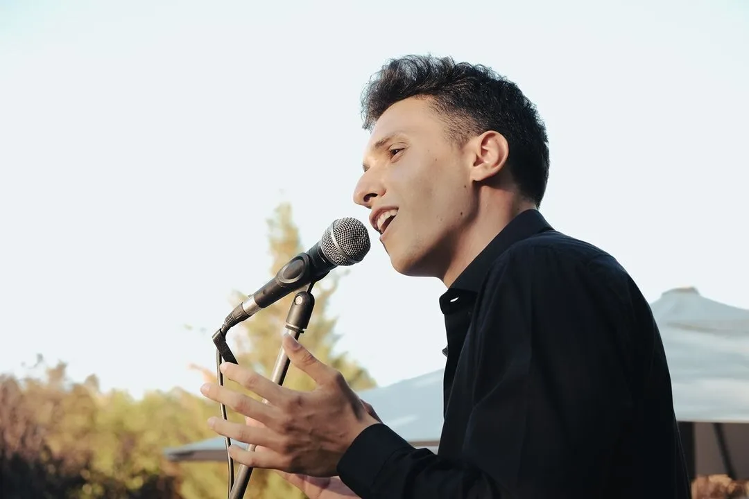 Primer plano de un cantante de perfil, con camisa negra, cantando en un micrófono sobre un soporte. Tiene una mano levantada mientras interpreta, y al fondo se ven árboles desenfocados y una estructura tipo carpa, un ambiente que encaja con pop español con flamenquito para bodas en Barcelona.