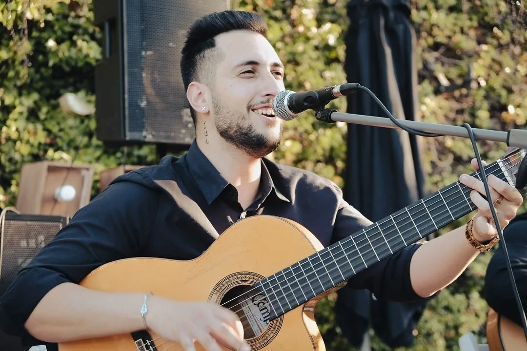 Primer plano de un músico sonriente cantando en un micrófono mientras toca una guitarra española; detrás se ve un altavoz y vegetación, una imagen asociada a pop español con flamenquito para bodas en Barcelona.